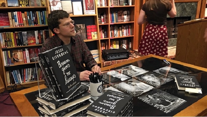 Jesse Eisenberg sitting in library in front a his books on the glass table